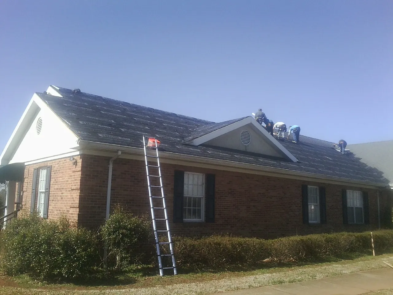 Crew tearing shingles off a brick ranch roof with a ladder set — Emergency Roof Repair work by our crew in Harmony