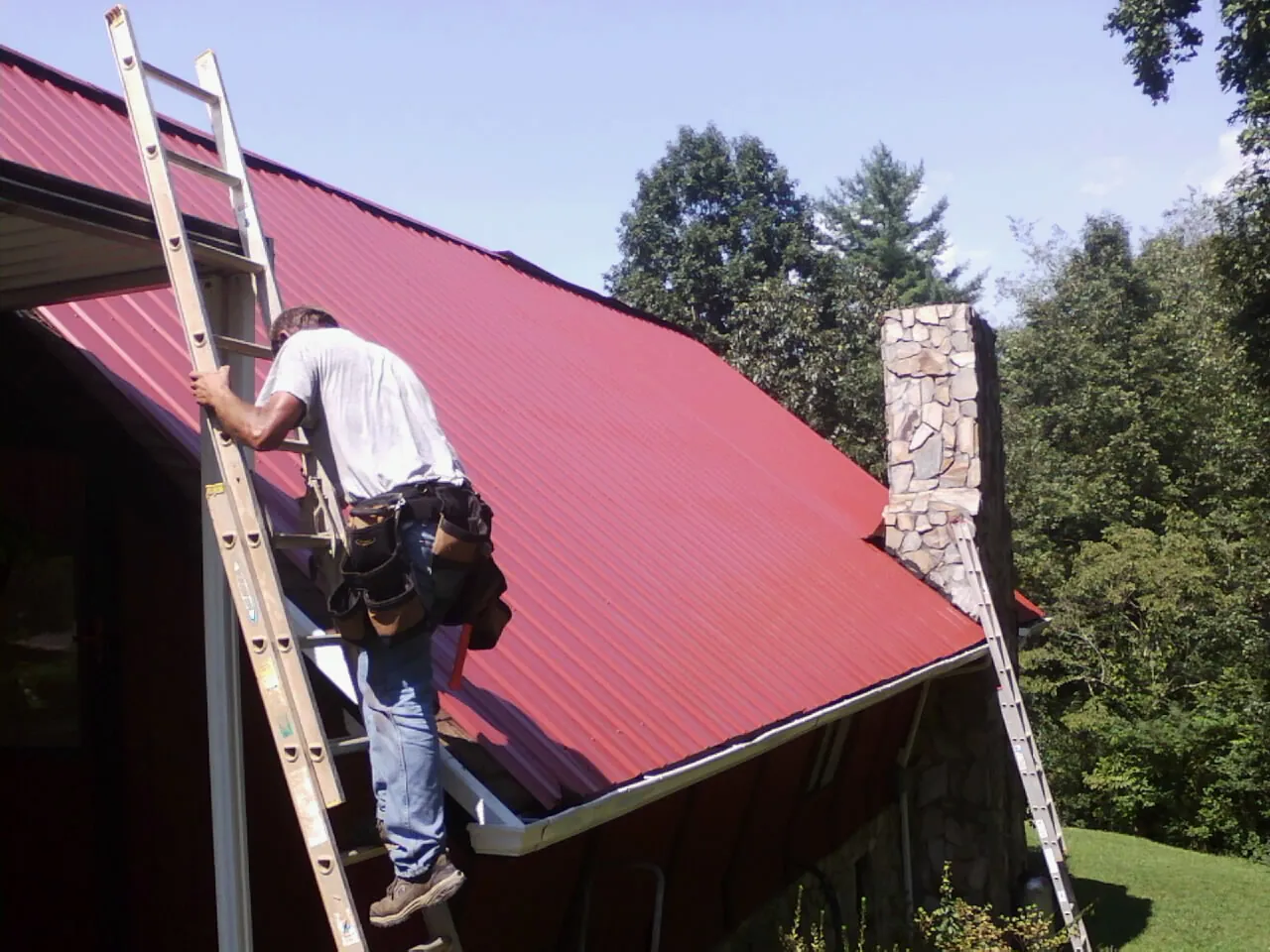Roofer on a ladder beside a red metal roof and stone chimney — Soffit & Fascia Repair by Roofing Brothers in Harmony